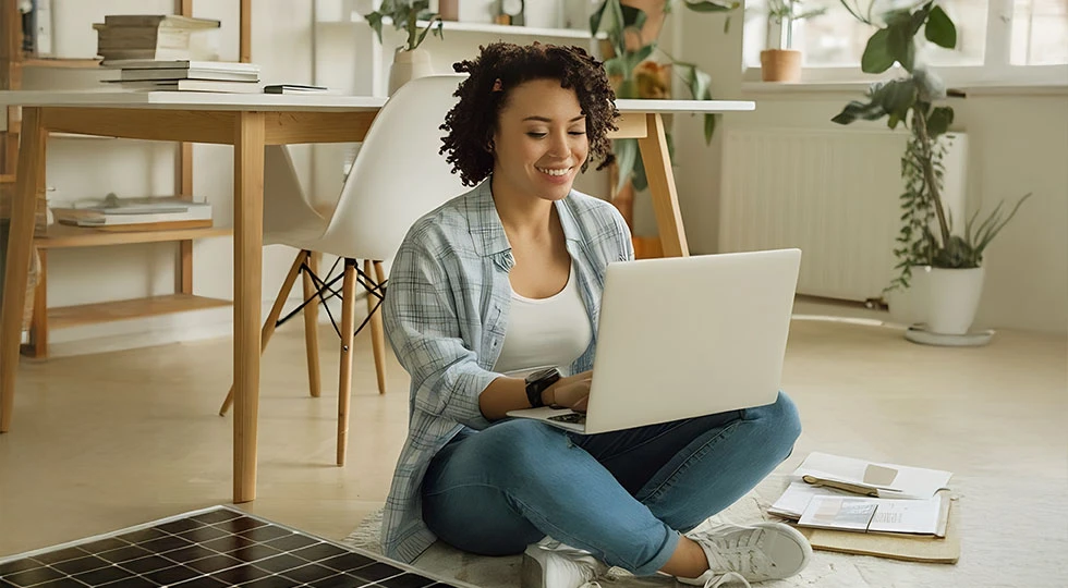 Jovem mulher usando laptop em ambiente de casa iluminado, sentada no chão com livros ao redor, representando trabalho remoto ou estudos em casa.