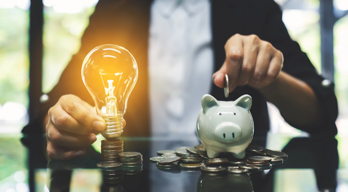 Pessoa colocando uma moeda na caixa de porquinho ao lado de uma lâmpada de ideia acesa, simbolizando economia e inovação financeira.