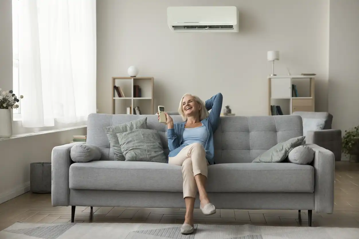 Mulher idosa sorridente relaxando no sofá enquanto segura um celular, em uma sala moderna e bem iluminada com decoração minimalista.